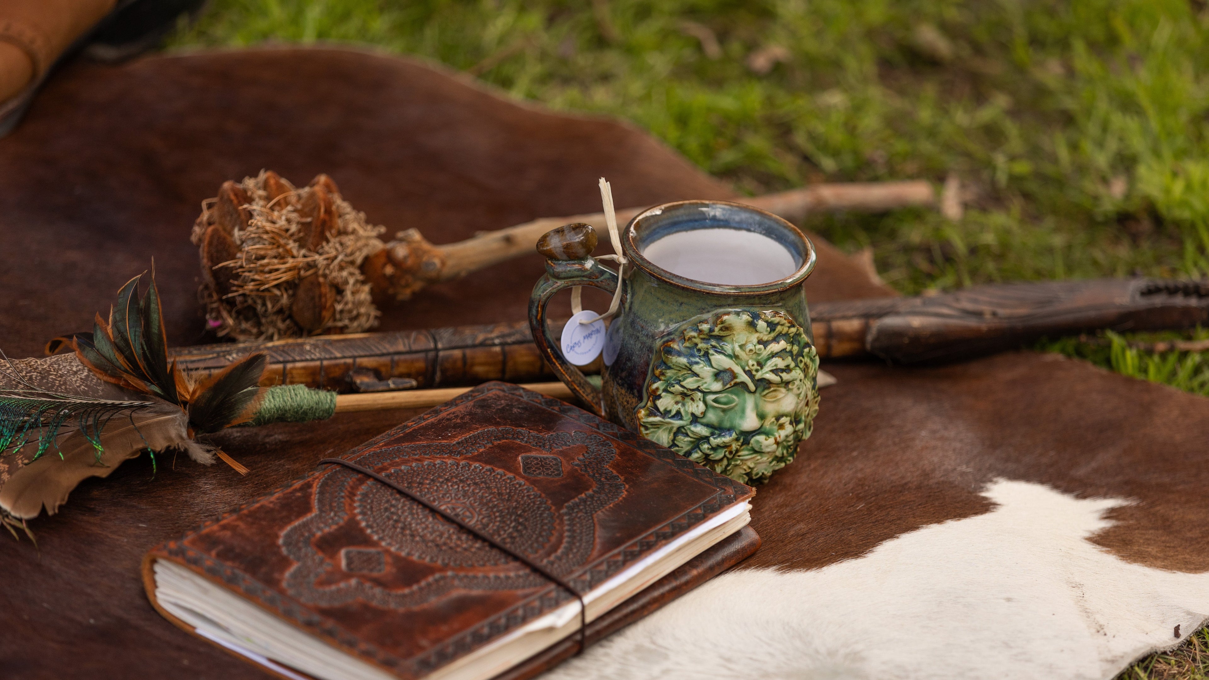 Leather journal, mug with greenery, and feather on a brown and white animal skin with grass in the background